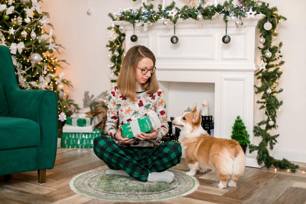 Junge Frau sitzt vor einem weihnachtlich geschmückten Kamin mit einem Geschenk im Schoß daneben ein Welsh Corgie. Beschriftung lautet: An Weihnachten freuen sich nicht nur Hundebesitzer sondern auch Hunde über Geschenke.